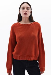 Overmaat, geribbelde gebreide trui in fel oranje, met een brede halslijn en lange mouwen, gecombineerd met donkere spijkerbroeken voor contrast.