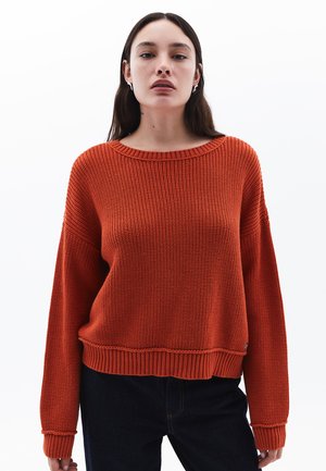 Overmaat, geribbelde gebreide trui in fel oranje, met een brede halslijn en lange mouwen, gecombineerd met donkere spijkerbroeken voor contrast.