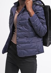 Námořnický modrý zateplený kabát s knoflíkovým zapínáním, prošívaným designem a límcem. V kombinaci s černou kabelkou a béžovým topem.