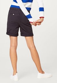 Shorts noirs pliés avec poches arrière, portés avec un pull à rayures bleu et blanc et des chaussures blanches à enfiler. Longueur visible jusqu'à la cheville.