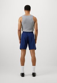 Top de algodão cinza sem mangas com calções azuis, apresentando uma cintura elástica. O modelo está de costas, com meias brancas e sapatos pretos.