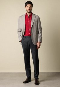 Man in een beige blazer, rode button-up overhemd, donkere broek en donkerbruine schoenen, staand tegen een effen beige muur.