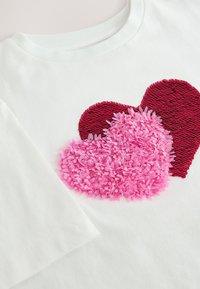 White cotton t-shirt featuring a red sequined heart and a pink textured heart, both prominently positioned on the front. Short sleeves.