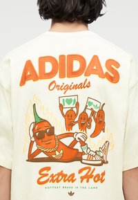 Πίσω μέρος κρεμ πουκάμισου με μεγάλο πορτοκαλί κείμενο "ADIDAS Originals" και καρτούν καυτερές πιπεριές που κρατούν πλακάτ, φλόγες και το σύνθημα "Extra Hot".