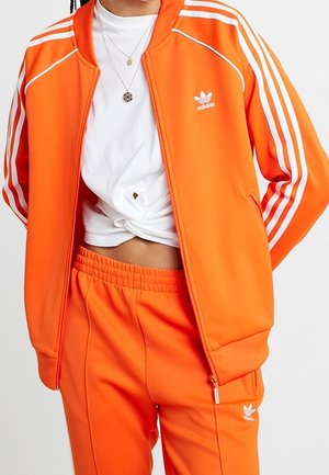 Person wearing bright orange Adidas tracksuit with white stripes and logo, paired with a white knotted crop top and layered necklaces.