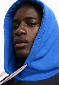 Close-up of a face wearing a blue hooded sweatshirt, featuring a smooth texture, drawstrings, and a dark-colored body. Neutral background.