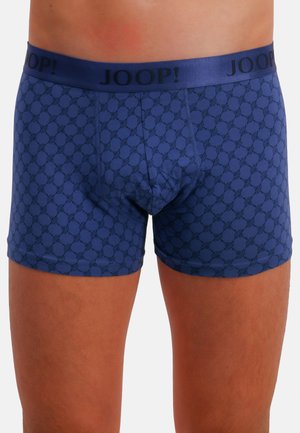 Blauwe jongensboxershorts met een herhalend cirkelpatroon en een zwart JOOP!-logo op de tailleband, gedragen door een staande persoon.