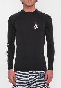 Czarny rash guard z długim rękawem wykonany z elastycznego materiału, z białym emblematem na klatce piersiowej i napisem wzdłuż rękawa, w połączeniu z paskowanymi szortami.