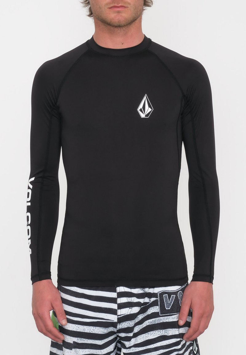 Czarny rash guard z długim rękawem wykonany z elastycznego materiału, z białym emblematem na klatce piersiowej i napisem wzdłuż rękawa, w połączeniu z paskowanymi szortami.