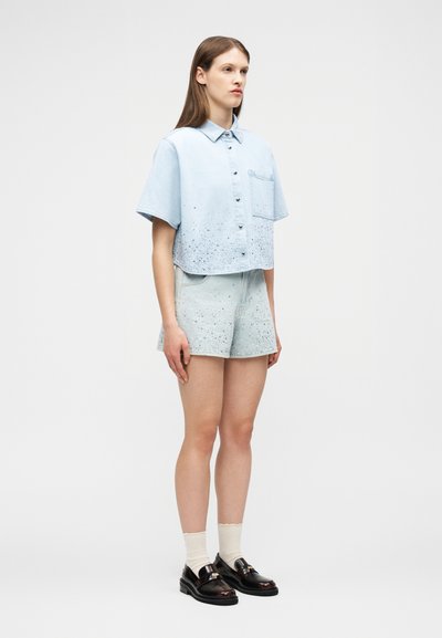 Γυναίκα που φοράει γαλάζιο κοντομάνικο πουκάμισο με κουμπιά και βουτυρωτό σχέδιο, ταιριαστά σορτσάκια, λευκές κάλτσες και μαύρα loafers, στέκεται μπροστά σε ουδέτερο φόντο.