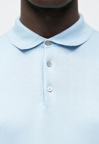 Gaiši zils polo krekls no adīta auduma, ar klasisku apkakli un trīs pogu šķēlumu, ar gludu tekstūru un vienkāršu dizainu.