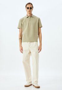 Polo shirt verde a texture, jeans beige e sneaker bianchi con dettagli marroni. Gli occhiali da sole completano l'outfit. Design semplice e moderno.