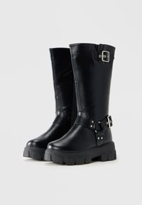 Botine din piele negre, până la jumătatea gambei, cu tălpi groase și bretele cu catarame argintii la gleznă și pe partea superioară, pe un fundal alb.