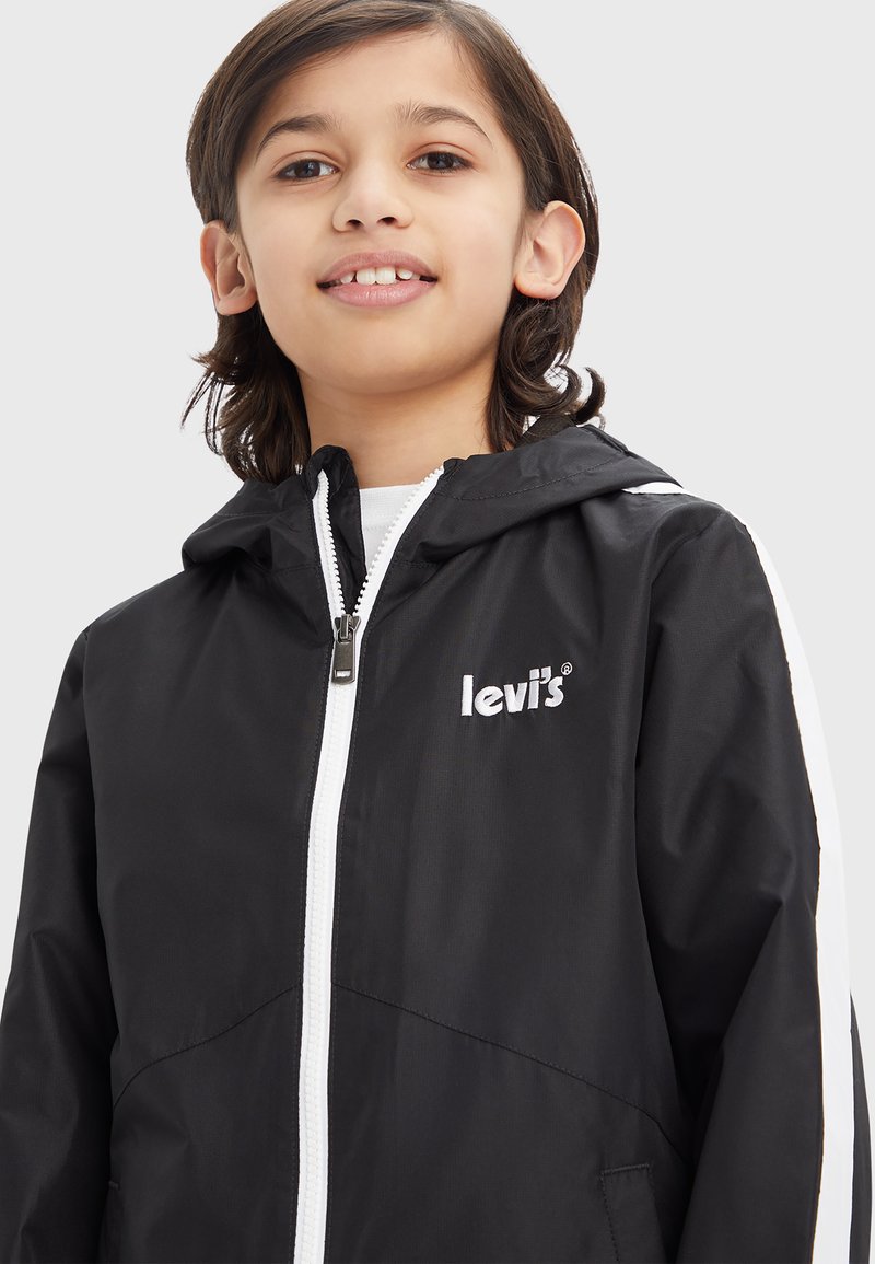 Schwarze Levi's Jacke mit Reißverschluss vorne, Kapuze, weißen Seitenstreifen und gesticktem Logo. Das Material wirkt leicht und hat eine glatte Textur.
