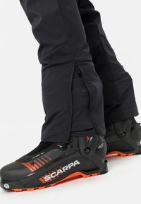 Des pantalons de ski noirs avec des zips sur les côtés et une texture lisse, associés à des bottes de ski noires et orange présentant des semelles robustes et un ajustement confortable.