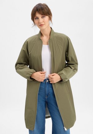 Woman wearing a long olive green zip-up jacket over a white top and blue jeans, holding jacket sides near the waist.