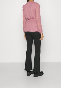 Blouse à manches longues rose pâle texturée avec taille à cordon, associée à un pantalon noir évasé et des chaussures noires épaisses.