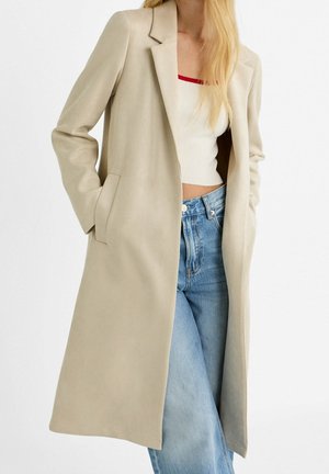 Blonde woman wearing a beige long coat over a white cropped ribbed top with red trim and light blue high-waisted jeans, hands in pockets.