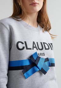 Szara bluza z napisem "CLAUDI" i "PARIS" w czarnym kolorze, z ozdobną kokardą w paski w odcieniach niebieskiego i granatowego z przodu. Miękka tekstura tkaniny.
