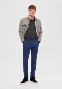 Siva flanel košulja na dugih rukava, tamna majica, plave hlače, crne tenisice s bijelim potplatima. Ležerni outfit s opuštenim krojem.