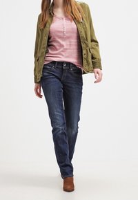 Olivově zelená bomber bunda přes červený pruhovaný henley tričko, doplněná o tmavě modré skinny džíny a hnědé kotníčkové boty.