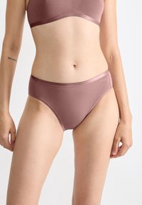 Vrouw in mauve naadloze hoge onderbroek en bijpassende beha met een zichtbare Romeinse cijfer tatoeage op haar linker onderarm.