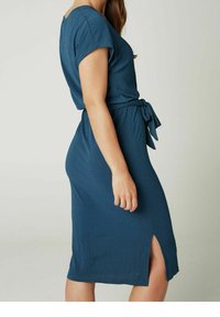 Vestido teal a la altura de la rodilla en tejido texturizado. Mangas cortas, cinturilla con lazo y detalle de abertura lateral. Diseño simple, ajustado, con una silueta relajada.
