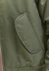 Maslinasto zeleni bomber jakna s glatkom, sjajnom teksturom, koja sadrži veliki, zaobljeni zakrpa džep s lijeve strane i rebraste rukave.