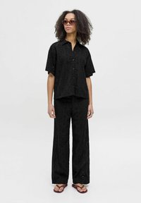 Chemise en coton noir à œillets et pantalon large assorti, présentant des découpes florales, une fermeture à boutons et une coupe décontractée. Porté avec des tongs.