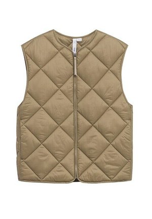 Person, die eine beige gesteppte Weste über einem beigen Pullover, weiße zerrissene Jeans und schwarze Knöchelstiefel trägt, steht vor einem einfarbigen Hintergrund.