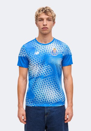 Jeune homme portant un maillot de football bleu et blanc à motif géométrique avec le logo New Balance et l'emblème du FC Porto, debout devant un fond uni.