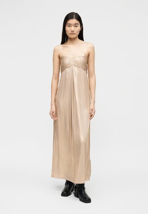 Žena s dlouhými tmavými vlasy, která má na sobě lesklé béžové dlouhé šaty bez rukávů a černé kotníkové boty, stojící na jednoduchém světlém pozadí.