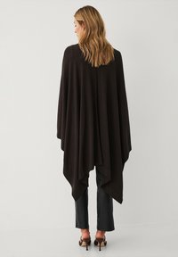 Brun, oversized poncho med en draperet design. Den bløde tekstur og den spidse sømkant skaber et lagdelt look. Bæres over sorte tætsiddende bukser og hæle.