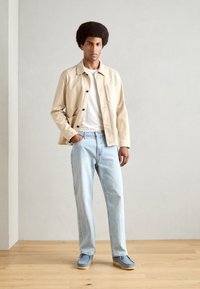 Lichtbeige knoopjacket, wit t-shirt, lichtblauwe jeans en blauwe schoenen. Gladde stof en comfortabele pasvorm. Eenvoudig zakdetail op de jas.