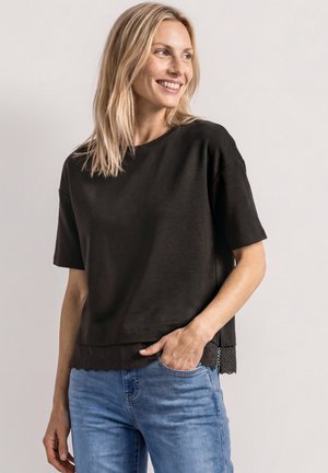 Kobieta o blond włosach uśmiecha się, spoglądając w bok, ubrana w czarny krótki top z koronkowym brzegiem i niebieskie dżinsy, ręka w kieszeni.