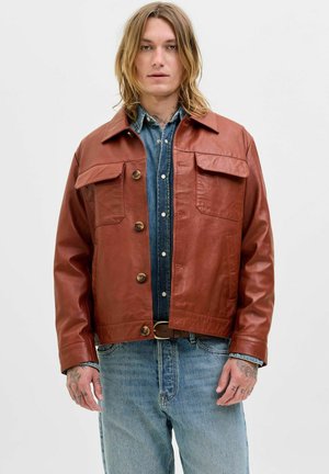 Chaqueta de cuero marrón con cuatro bolsillos, cierre de botones y un diseño ligeramente corto. Lleva puesta una camisa de mezclilla y unos jeans de color azul claro.