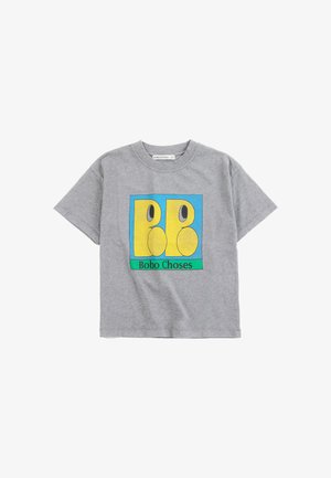 Garçon aux cheveux foncés portant un T-shirt gris avec un logo coloré jaune et bleu de Bobo Choses et un short vert et violet.