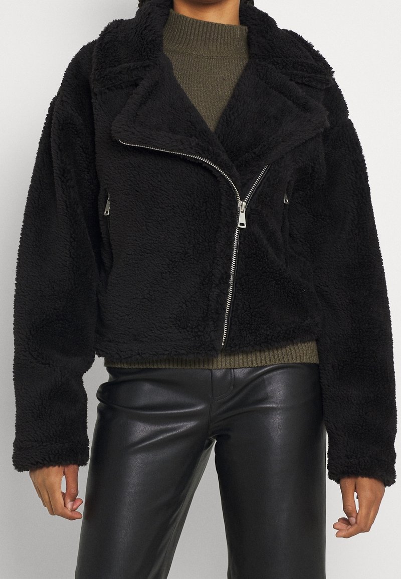 Femme portant une veste zippée noire en tissu duveteux par-dessus un pull vert et un pantalon en cuir noir, debout devant un fond uni.