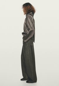 Osoba s kudrnatými vlasy nosí volný, lesklý černý top s vysokým límečkem a širokými tmavými kalhotami, stojí bokem s rukama v kapsách.