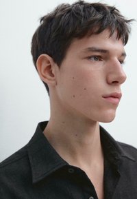 Ung mand med kort mørkt hår iført en sort skjorte med krave, der kigger til højre mod en lys, ensfarvet baggrund.