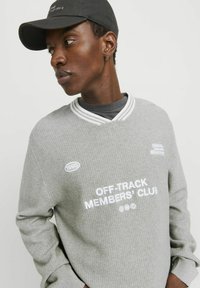 A szürke waffle-kötésű pulóver fehér V-nyakú gallérral rendelkezik, és hímzett felirata van: "OFF-TRACK MEMBERS' CLUB." Tartalmaz egy kis kerek foltot, amelyen a "1990" felirat olvasható.
