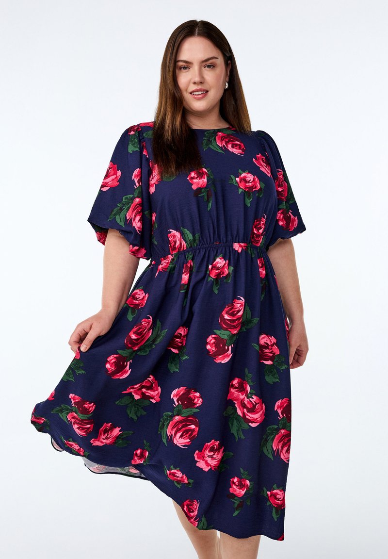 Bloemenjurk in donkerblauwe stof, met grote roze rozen en groene bladeren. Losse pasvorm met een geraffineerde taille en korte, brede mouwen.
