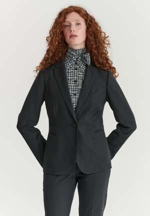 Femme aux longs cheveux roux bouclés portant un costume gris foncé et un chemisier à motif noir et blanc avec col montant, debout les mains derrière le dos.