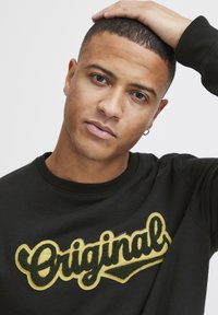Sudadera negra con bordado texturizado en amarillo que dice "Original", con cuello redondo y puños de canalé, llevada por una persona con cabello corto.