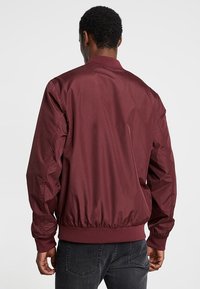 Burgundzka bomberka wykonana z gładkiego materiału, z elastycznymi mankietami i pasem w talii; charakteryzuje się prostym fasonem bez wyraźnych wzorów czy detali.