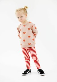 Világos rózsaszín pulóver narancssárga szívmintákkal, piros-fehér csíkos leggings-szel és fekete sportcipővel, fehér akcentussal párosítva.