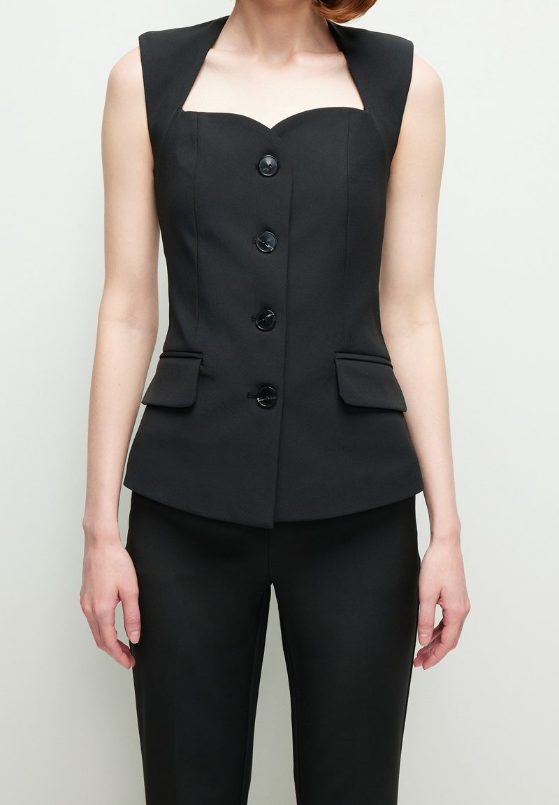 Femme portant un gilet noir sans manches, ajusté, à boutons et poches à rabat, avec un pantalon noir assorti, debout devant un fond uni.