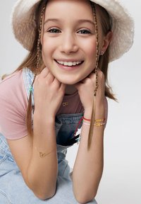 Dziewczyna z jasnobrązowymi włosami w warkoczu, nosząca biały kapelusz typu bucket, różową koszulkę i dżinsowe ogrodniczki, prezentująca złotą bransoletkę w kształcie serca oraz kolorowe akcesoria na nadgarstku.