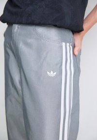 Grijze sportbroek met drie witte strepen langs de zijkanten, met een klein wit Adidas-logo op het voorzakgebied en een stijlvolle afwerking.