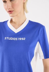 Sininen ja valkoinen lyhythihainen t-paita V-päällä, jossa edessä on valkoisella tekstillä painettu "STUDIOS 1992". Pehmeää puuvillakangasta.